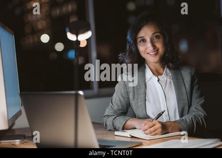 Portrait of a smiling young businesswoman sitting at her office desk writing notes and using a laptop while working overtime at night Stock Photo
