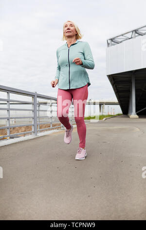 Active mature blonde female runner in sportswear exercising along bridge border while moving forwards in urban environment Stock Photo