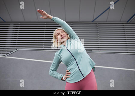 Mature active female in sportswear doing side-bends under bridge construction while spending time in urban environment Stock Photo