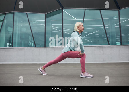 Healthy and active mature blonde sportswoman exercising by contemporary architectural building in urban environment Stock Photo