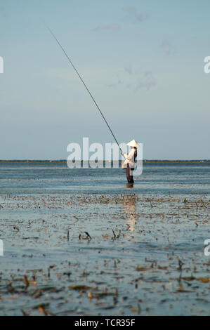 Typical Balinese fisherman is standing in the shallow water while he is checking his bite of the fishing rod at Sanur Beach in Bali, Indonesia Stock Photo