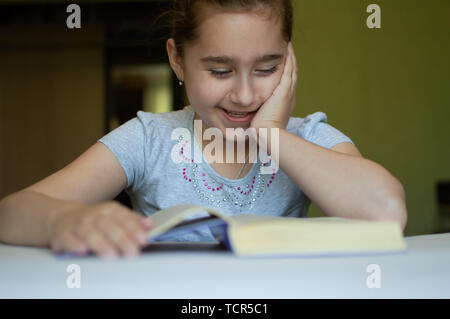 tortured girl child reads a book at the table and she is very tired and wants to sleep, yawns, falls asleep while reading stories and lessons Stock Photo