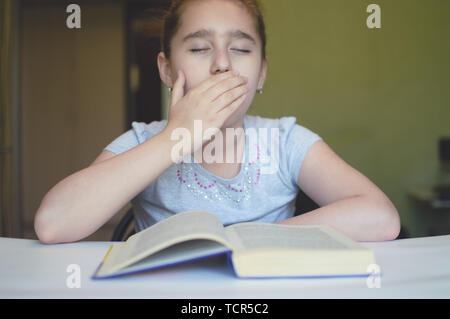 tortured girl child reads a book at the table and she is very tired and wants to sleep, yawns, falls asleep while reading stories and lessons Stock Photo