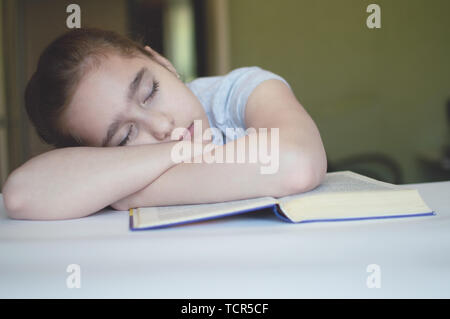 tortured girl child reads a book at the table and she is very tired and wants to sleep, yawns, falls asleep while reading stories and lessons Stock Photo