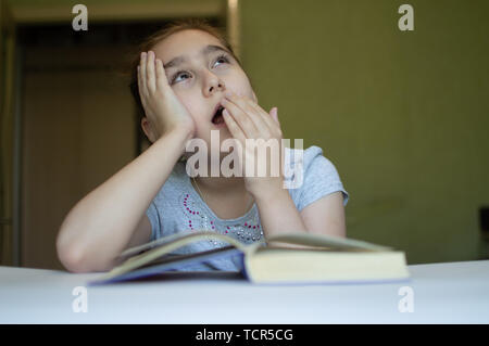 tortured girl child reads a book at the table and she is very tired and wants to sleep, yawns, falls asleep while reading stories and lessons Stock Photo