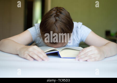 tortured girl child reads a book at the table and she is very tired and wants to sleep, yawns, falls asleep while reading stories and lessons Stock Photo