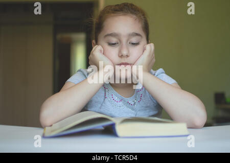tortured girl child reads a book at the table and she is very tired and wants to sleep, yawns, falls asleep while reading stories and lessons Stock Photo