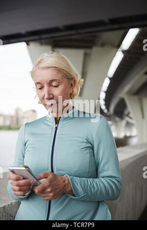 Contemporary active blonde mature female in sportswear scrolling in smartphone in urban environment Stock Photo