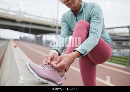 Mature contemporary woman in activewear tying shoelace of sneaker while standing on racetrack in urban environment Stock Photo