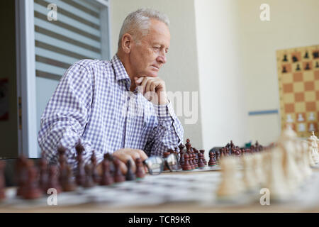 Old man in checked shirt holding glasses and thinking about next chess move  in game with imaginary opponent Stock Photo