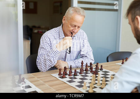 Focused old man in checked shirt biting his glasses while playing chess with his young son Stock Photo