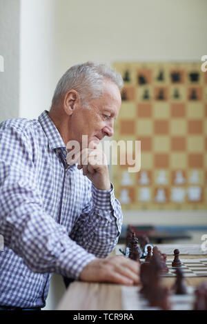 Old man in checked shirt with silver hair looking with smile at chess board and analyzing interesting chess game Stock Photo