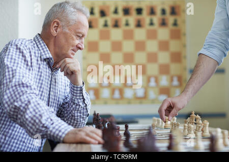 Old man in checked shirt holding glasses and thinking about next chess move  in game with imaginary opponent Stock Photo