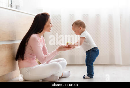 Mother giving fresh apple to baby, sitting on floor in kitchen