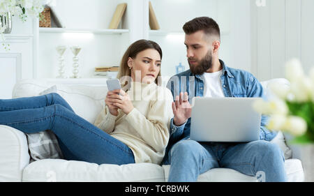 Woman Spying Husband's Laptop, Sitting Together On Sofa Stock Photo