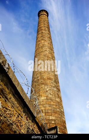 Razor wire on top of stone wall with old factory mill chimney against sky in the background Stock Photo