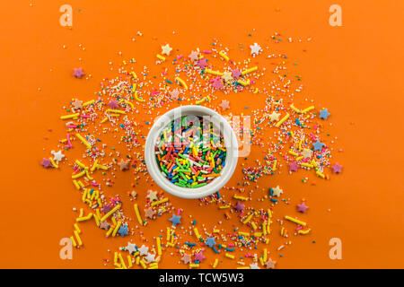 Sprinkles on bright orange background with little white bowl in middle of background and multi-coloured sprinkles sprayed around Stock Photo
