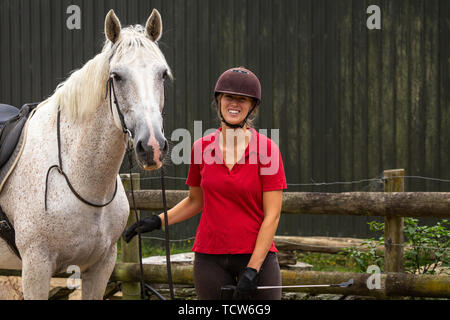 An attractive happy smiling young woman dressed in a red polo shirt with her white horse ready for a ride, copyspace to the right Stock Photo