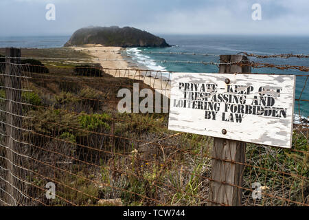 A worn no trespassing sign in front of a fence topped with barbed wire, in the background a beautiful sandy beach and the crashing ocean. Stock Photo