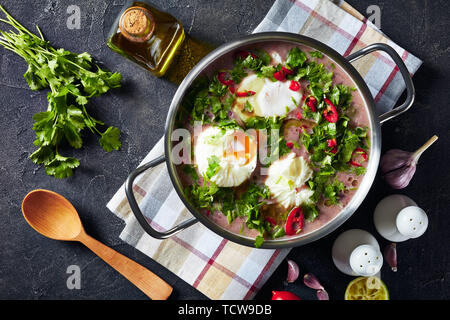 Fresh Homemade red kidney Bean Soup with poached eggs and cilantro in a metal pan on a concrete table with a wooden spoon and ingredients, view from a Stock Photo