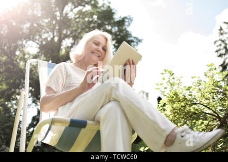Senior blonde woman sitting in chair and using digital tablet in garden Stock Photo