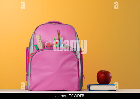 Student set. Pink backpack with office supplies, a book, a red apple on the table on a yellow background. Back to school. Stock Photo