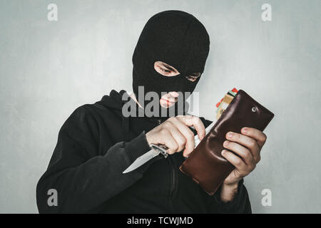 Masked criminal at the black background grins looking at the viewer and showing a purse in hand. The robber in the Balaclava counts the money in the Stock Photo