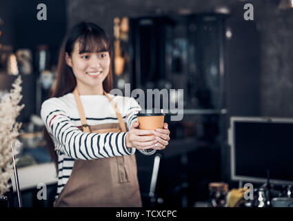 Asian Barista served coffee up with smile at cafe holding mug with two hand with smiling face at cafe bar background Stock Photo