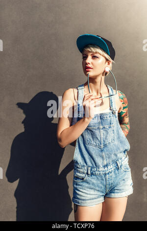 Young alternative girl wearing cap standing isolated on grey wall on the city street holding sunglasses looking aside thoughtful Stock Photo