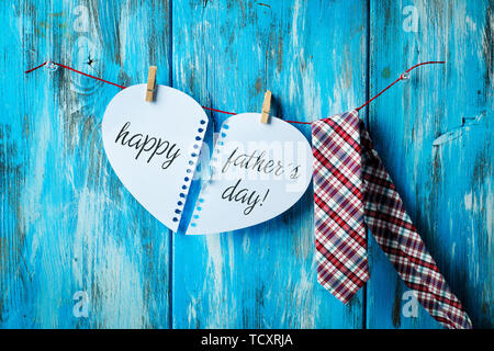 the text happy fathers day written in two notepad pages forming a heart hung with clothespins in a clothes line next to a necktie, against a rustic bl Stock Photo