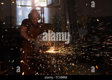 Concentrated professional blacksmith in ear protectors using cutting machine while working with metal in workshop Stock Photo