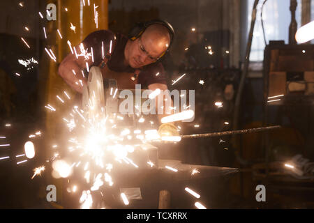 Concentrated young male blacksmith in ear protectors and safety goggles standing at anvil and cutting metal bar with powerful circular saw Stock Photo