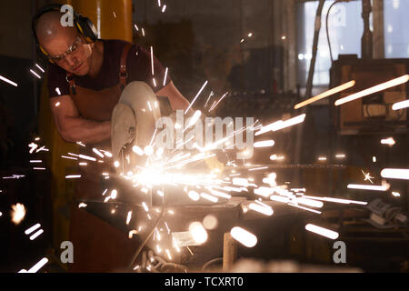 Serious busy bald metalworker in ear protectors standing by anvil and cutting metal with circular saw Stock Photo