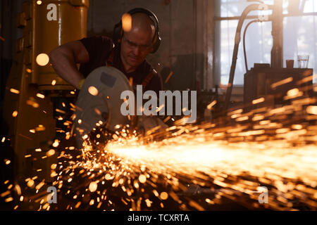 Serious skilled male blacksmith in ear protectors and safety goggles standing in smithy and using circular saw while cutting metal piece Stock Photo