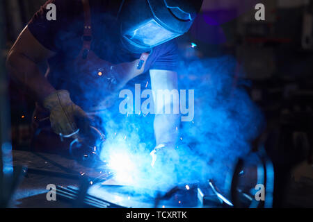 Busy mechanic in work helmet standing at desk and using welding torch while working with metal in smithy Stock Photo