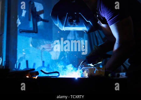 Busy factory worker in protective mask using torch while welding wrought iron in dark workshop, blue light from welding process Stock Photo