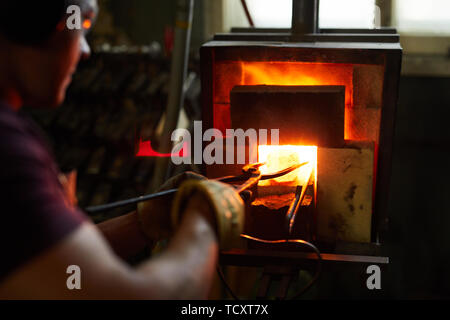 Over shoulder view of busy experienced blacksmith using tongs while putting metal piece into heated furnace Stock Photo