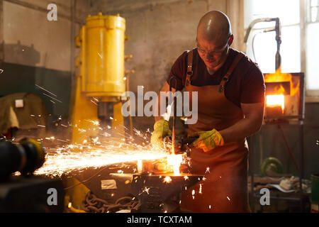 Frowning bald blacksmith in apron cutting heated metal piece with grinder on anvil while working in smithy Stock Photo