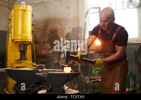 Strong young man in safety goggles and apron using sledgehammer to shape metal piece while working in smithy Stock Photo