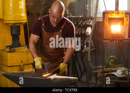 Focused young man in leather apron and protective gloves standing at anvil and beating hot iron with hammer Stock Photo