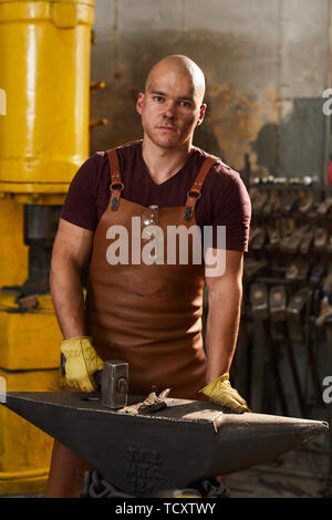 Portrait of serious strong bald young metalworker in brown apron standing at anvil and using hammer in workshop Stock Photo