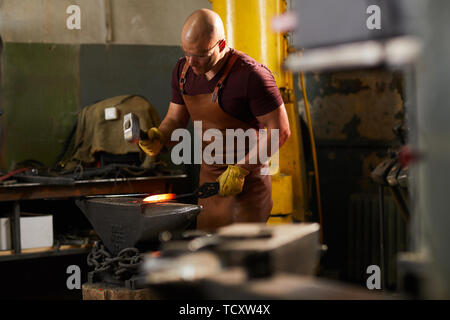 Focused young bald blacksmith in leather apron and protective goggles standing at anvil and forging hot metal with hammer Stock Photo