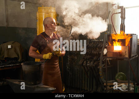 Serious bald young male metalworker in leather apron blowing on forged knife in smoke while checking it after cooling Stock Photo