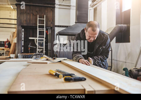 Serious joiner grinding timber with help of special equipment. Busy carpenter in uniform working with wood. Stock Photo