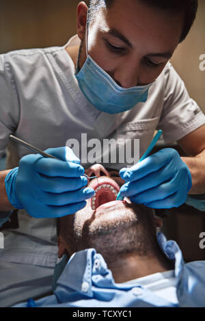 Cropped view of male dentist wearing white uniform, checking up patient's teeth using mouth mirror and dental explorer. Dental tools. Treatment. Moder Stock Photo