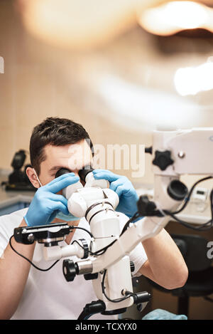 Male dentist wearing white uniform, looking into professional dental endodontic binocular microscope with camera in the office. Close up Stock Photo