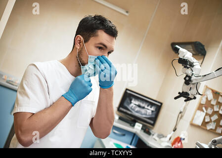 Young male dentist putting on a face mask and wearing blue rubber gloves while getting ready for an appointment with a patient. Cropped image Stock Photo