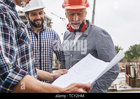 Project discussing. Senior engineer in formal wear and helmet looking at construction drawing with two young cheerful builders while standing against  Stock Photo
