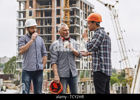 Lunch time. Group of builders in working uniform are eating sandwiches and talking while standing at construction site. Building concept. Taking a bre Stock Photo