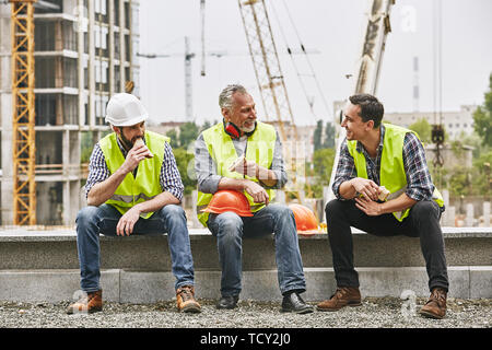 Time for a break. Group of builders in working uniform are eating sandwiches and talking while sitting on stone surface against construction site. Bui Stock Photo